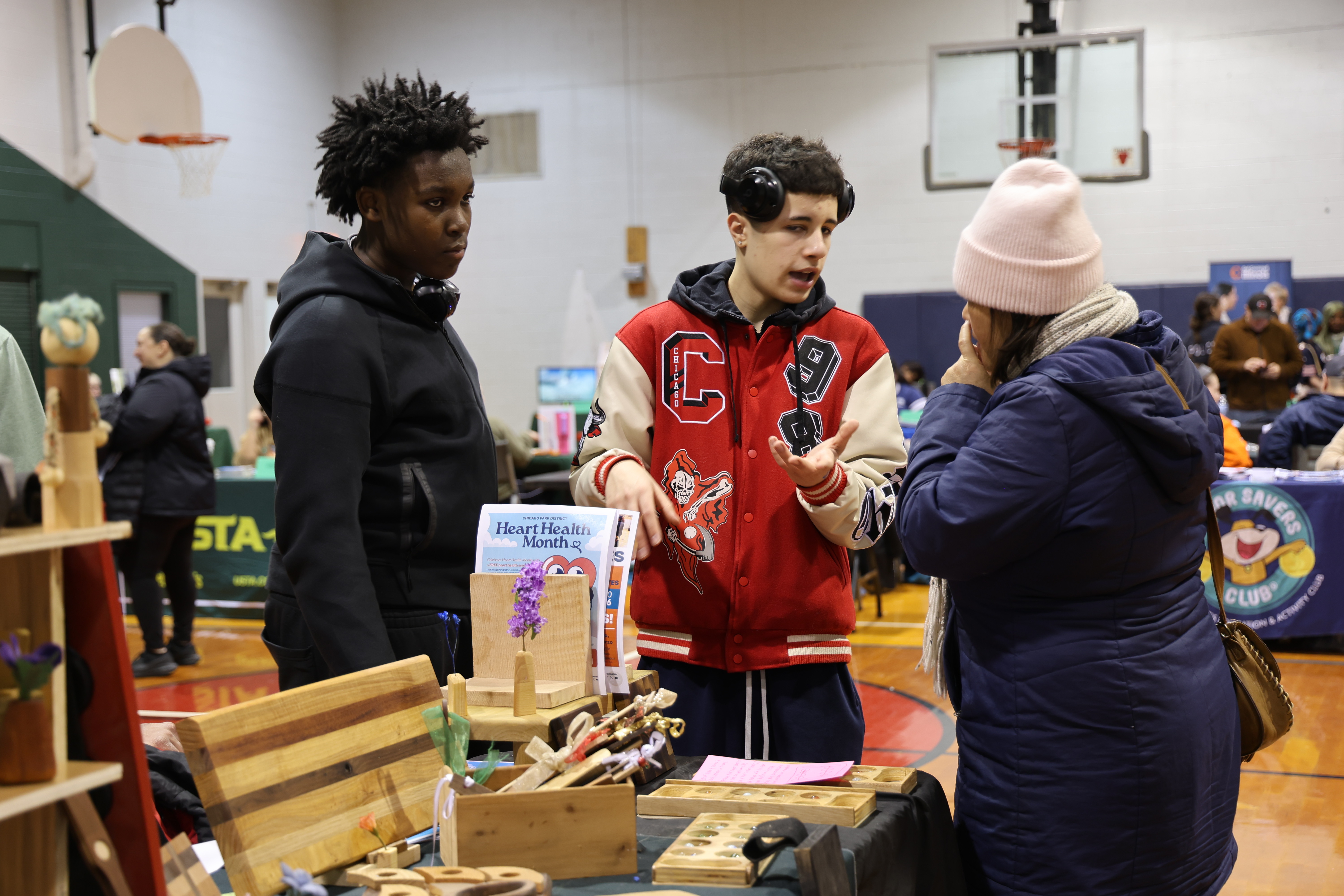 Three people talk at a craft table with wooden items in a gymnasium.